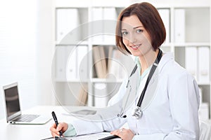 Young brunette female doctor sitting at the table and working at hospital office.