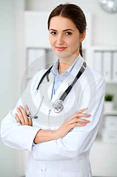 Young brunette female doctor standing with arms crossed and smiling at hospital. Physician ready to examine patient
