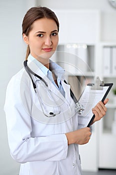 Young brunette female doctor standing with clipboard and smiling at hospital. Physician ready to examine patient