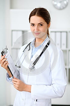 Young brunette female doctor standing with clipboard and smiling at hospital. Physician ready to examine patient