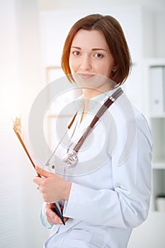 Young brunette female doctor standing with clipboard and smiling in hospital