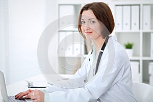 Young brunette female doctor typing on laptop comoputer while sitting at the table in hospital office. Health ca