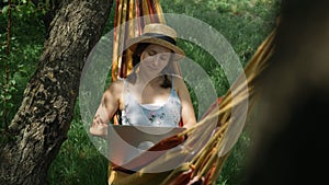 Young brunette female in hat lying in hammock and working on laptop at green garden. Charming woman resting in hammock and using l