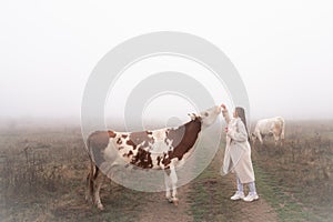 Young brunette girl in coat stands among the medow in fog with cow that reaching to her hand