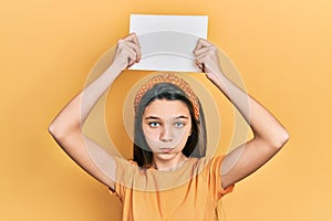 Young brunette girl holding blank empty banner over head puffing cheeks with funny face