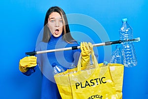Young brunette girl holding recycling bag with plastic bottles and waste picker afraid and shocked with surprise and amazed