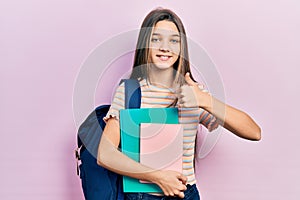 Young brunette girl holding student backpack and books smiling happy and positive, thumb up doing excellent and approval sign