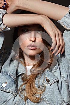 Young brunette girl with long flowing hair dressed in jeans jacket poses holding her hands on her head on the dark