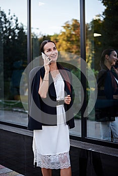 Young brunette girl with red pony tail, wearing stylish black jacket and white silk dress, standing near modern glass building,