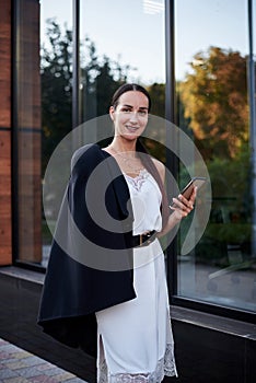 Young brunette girl with red pony tail, wearing stylish black jacket and white silk dress, standing near modern glass building,