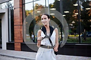 Young brunette girl with red pony tail, wearing stylish white silk dress and black blouse, standing in front of glass building.