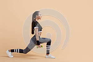 A young brunette girl shakes her gluteal muscles using extra weights, full-length photo, against a peach background.