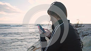 Young brunette girl sits on rocky beach and looks at photos in the phone