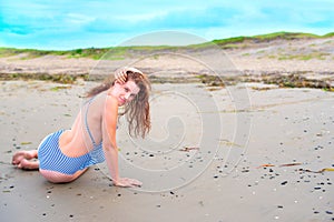 Young brunette girl in striped swimsuit sitting on the beach on the sand Shining bright sun blue sky clouds