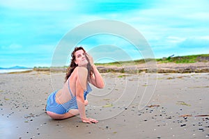 Young brunette girl in striped swimsuit sitting on the beach on the sand Shining bright sun blue sky clouds