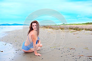 Young brunette girl in striped swimsuit sitting on the beach on the sand Shining bright sun blue sky clouds