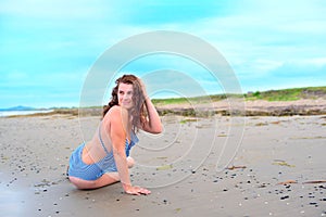 Young brunette girl in striped swimsuit sitting on the beach on the sand Shining bright sun blue sky clouds