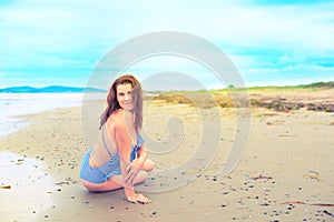 Young brunette girl in striped swimsuit sitting on the beach on the sand Shining bright sun blue sky clouds