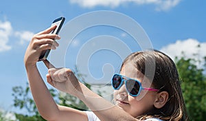 Young brunette girl in summer day enjoying the sun, making selfie portrait. Baby holds a mobile phone in hand and smiles