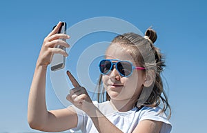 Young brunette girl in summer day enjoying the sun, making selfie portrait. Cute baby holds a mobile phone in hand and smiles