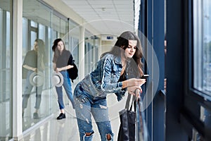 Young brunette girl, wearing casual jeans clothes, leaning on railing in light airport hallway, checking phone, with her