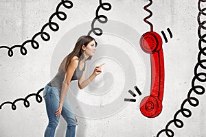 Young brunette girl wearing casual jeans and t-shirt pointing finger to big red cartoon telephone receiver drawn on