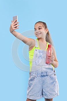 A young brunette girl wearing  denim overalls shorts stands with a phone and with a bottle of water
