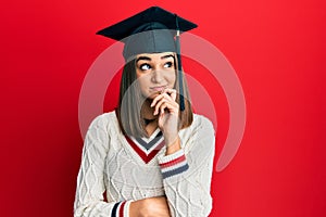 Young brunette girl wearing graduation cap serious face thinking about question with hand on chin, thoughtful about confusing idea