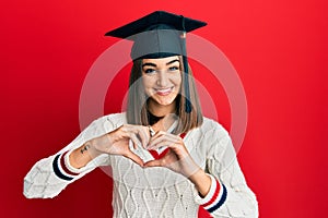 Young brunette girl wearing graduation cap smiling in love showing heart symbol and shape with hands