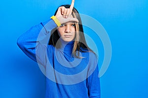 Young brunette girl wearing turtleneck sweater making fun of people with fingers on forehead doing loser gesture mocking and