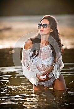 Young brunette girl in wet white blouse posing provocatively in water. Sensual attractive woman with black sunglasses, summer