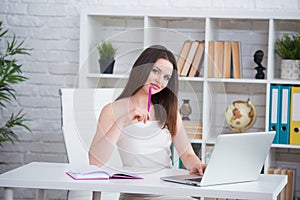 A young brunette girl in a white dress is sitting in the office at the table. Woman works at the laptop