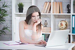 A young brunette girl in a white dress is sitting in the office at the table. Woman works at the laptop