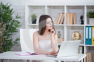 A young brunette girl in a white dress is sitting in the office at the table. Woman works at the laptop