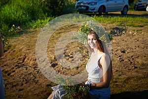 Young brunette girl in a white dress, sundress and a wreath of flowers in summer on the coast of river or lake in the