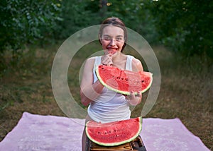 A young brunette girl in a white T-shirt with bright red lipstick on her lips poses with a huge slice of ripe watermelon in nature