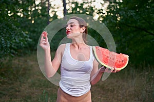 A young brunette girl in a white T-shirt with bright red lipstick on her lips poses with a huge slice of ripe watermelon in nature