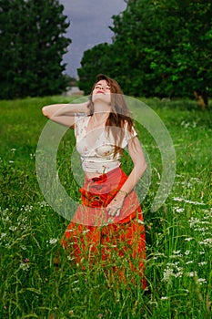 Young brunette girl in white top and red skirt standing in green meadow field grass in countryside looking up touching her hair