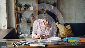 Young brunette in glasses in designing illustration cutting splash-paper then sticking figures in notebook using