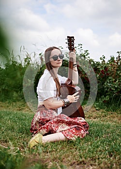 Young brunette hippie woman, wearing boho style clothes, sitting on green grass, holding guitar. Indie musician relaxing on green