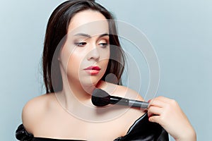 A young brunette looks to the side and holds a black makeup brush in front of her