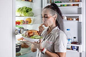 A young brunette makes a late night craving for roast chicken in front of the open fridge
