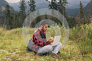 Young brunette male in jeans and shirt is working at a computer while being on the nature.