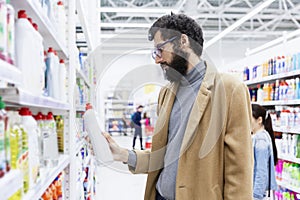 Young brunette man with glasses with a beard in a supermarket chooses a cleaning product for the house
