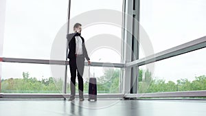 Young brunette man with suitcase on the wheels enters the room with panoramic windows while talking on the phone. Front