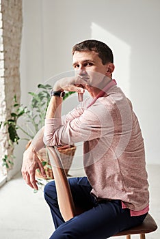 Young brunette man, wearing casual light pink jumper, sitting on brown stool in light room, thinking. Serious emotions.Three-
