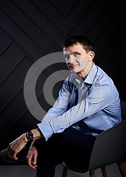 Young brunette man, wearing light blue shirt, sitting in grey armchair in front of black wall in photo studio. Businessman resume