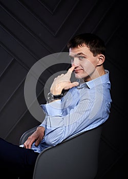 Young brunette man, wearing light blue shirt, sitting in grey armchair in front of black wall in photo studio, leaning on his chin