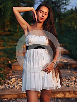 Young brunette model at the white dress staying on the stairs with green trees  with closed eyes