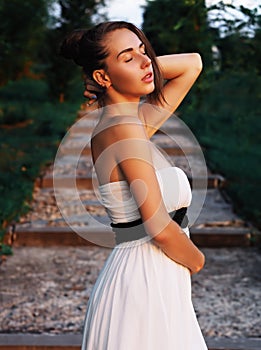 Young brunette model at the white dress staying at the stairs with green trees with closed eyes